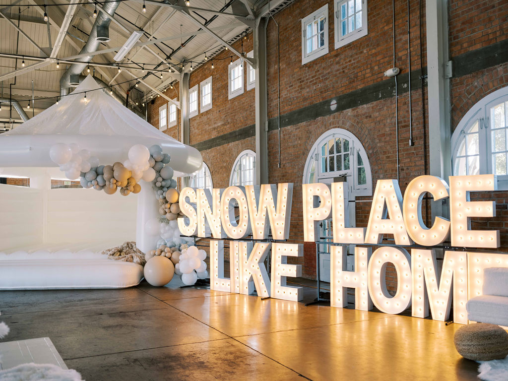 A company holiday party setup at BRICK in San Diego, California. Large letters with LED lighting spell out "Snow Place Like Home" next to a white bounce house.