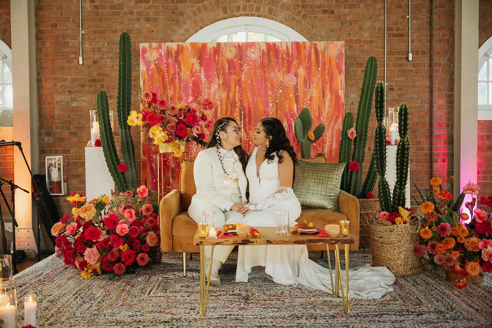 Two brides seated in front of a colorful wedding backdrop