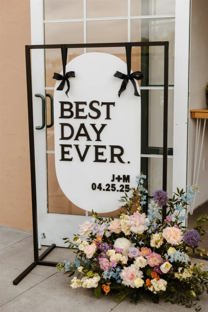 White wedding welcome sign reading “Best Day Ever” with the couple’s name and date, accented with black bows and a floral arrangement, at a San Diego LGBTQ+ wedding at BRICK